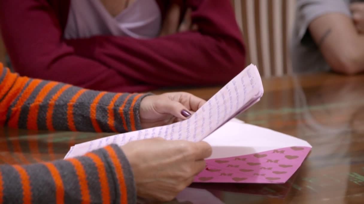 Someone in a striped orange and gray sweater is carefully unfolding a letter, the pink envelope decorated with hearts. The letter's contents are hidden, but the anticipation is palpable.
