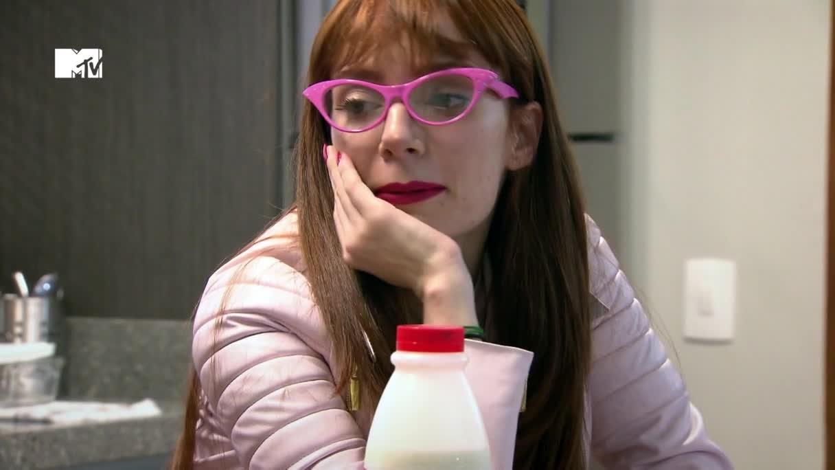 A woman with bright pink glasses rests her chin on her hand, looking thoughtful. A bottle of milk sits on the counter in front of her, and the MTV logo is visible in the corner.
