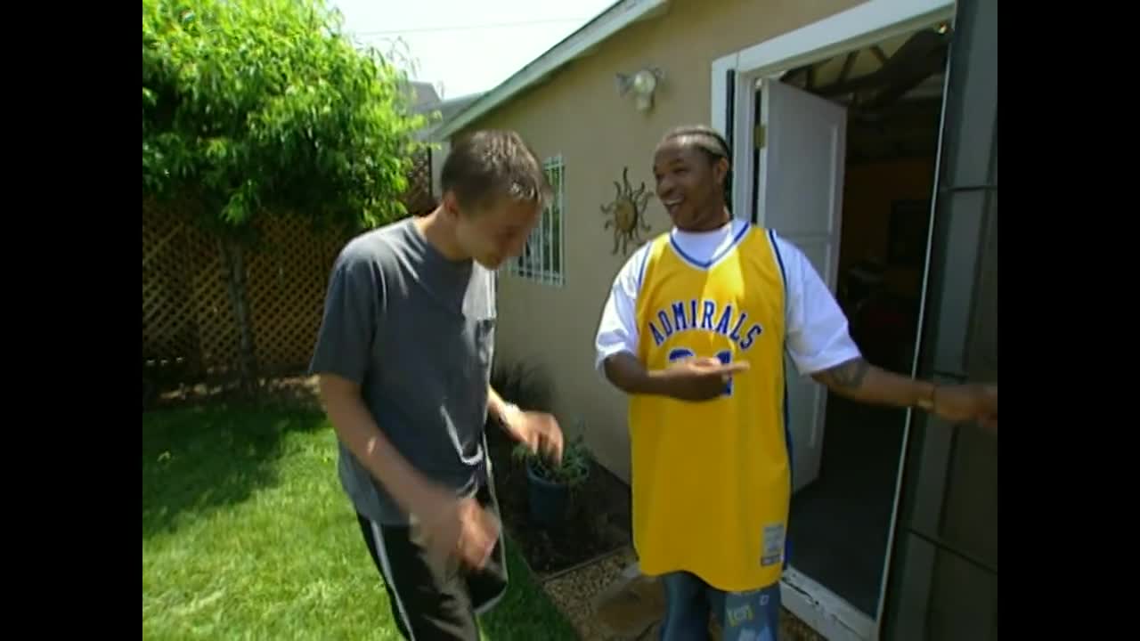 A man in a yellow jersey gestures towards the open door, while the other man, wearing a gray t-shirt, looks down and gestures with his hands. The pair stand in a sunlit backyard, near a small building.
