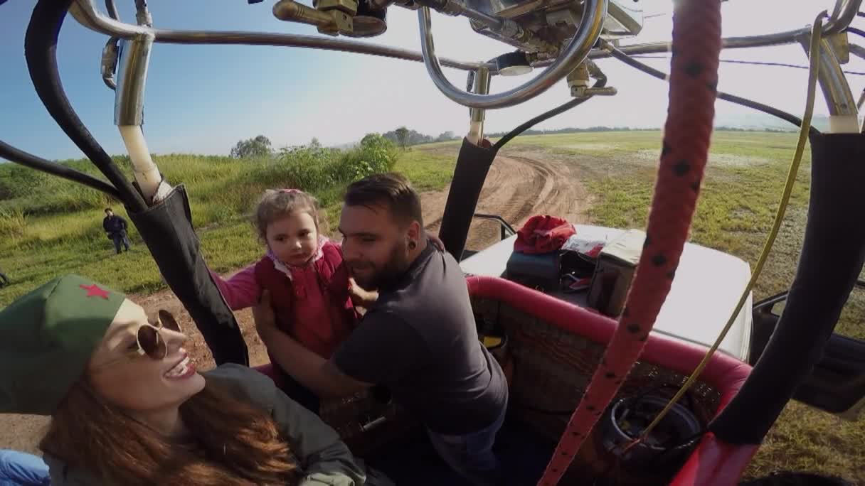 Inside a hot air balloon basket, a man holds a small child while a woman with a red star hat smiles. The balloon is rising over a grassy field, with a dirt road visible below.
