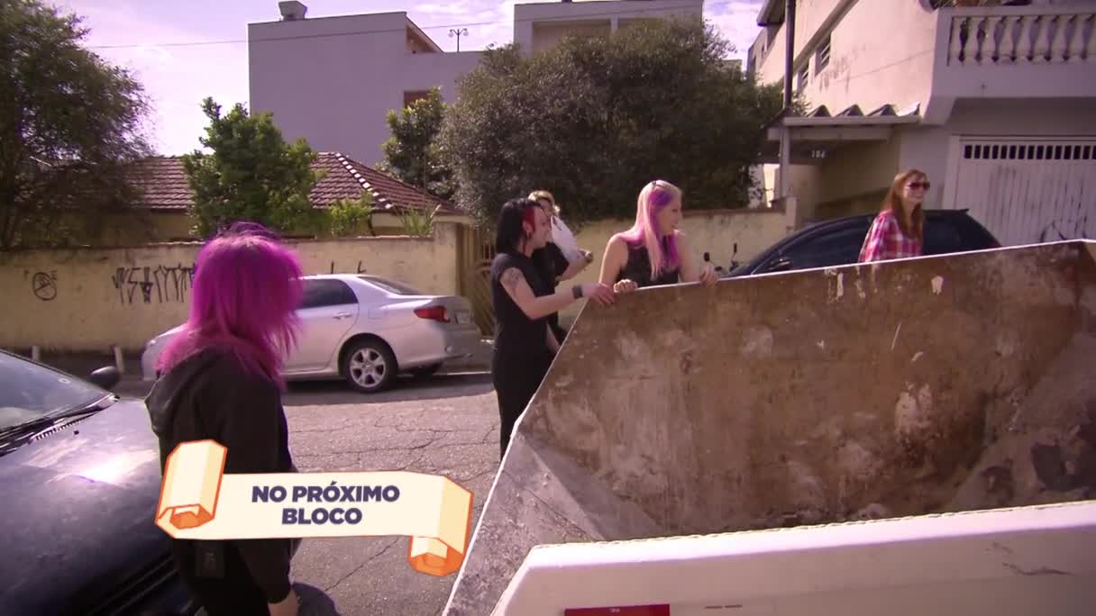 A group of young women are gathered around a large, open dumpster on a residential street in Brazil. One with bright pink hair stands near a car, while others lean against the edge of the container, looking onward.
