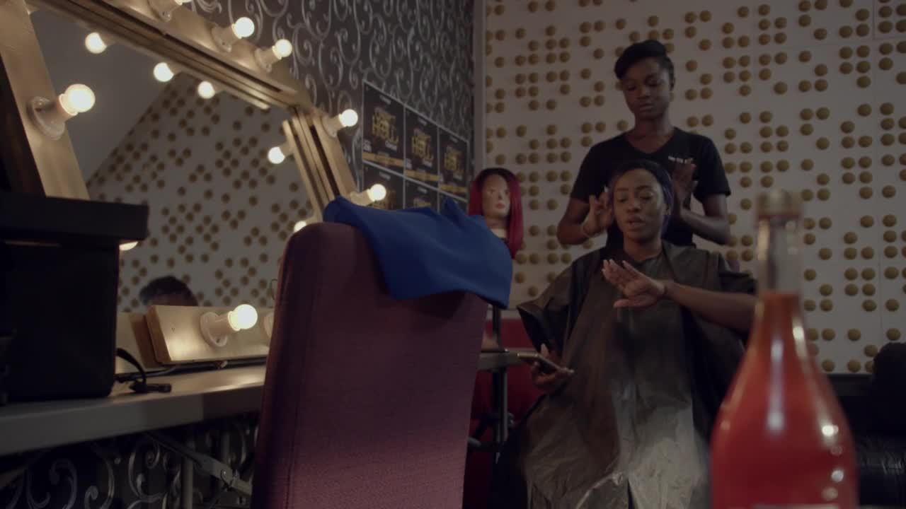 In a brightly lit room, a woman in a salon chair is getting her hair styled.  Her hands are up, and the stylist is working on her hair, probably for a shoot on MTV Brazil.

