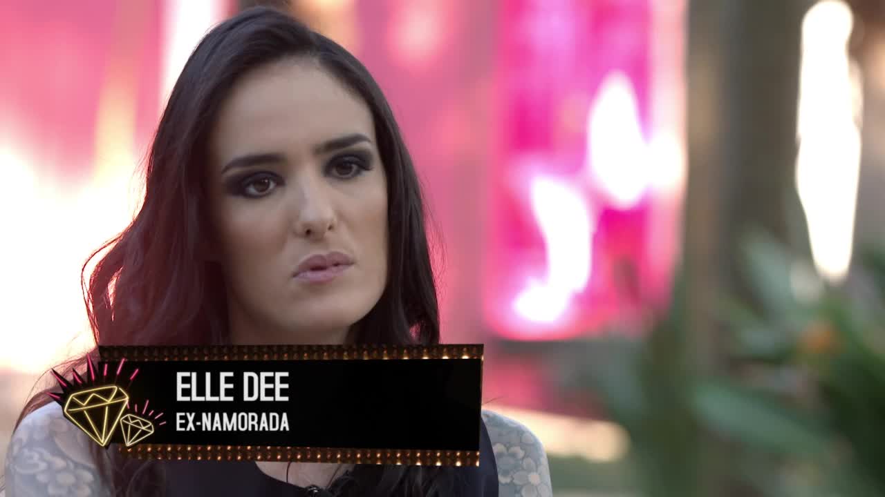 Elle Dee, identified as an ex-girlfriend, is speaking on camera, her face framed by dark hair and heavy eye makeup. The background is a soft, blurred mix of pink and light, suggesting a studio setting for an MTV Brazil show.
