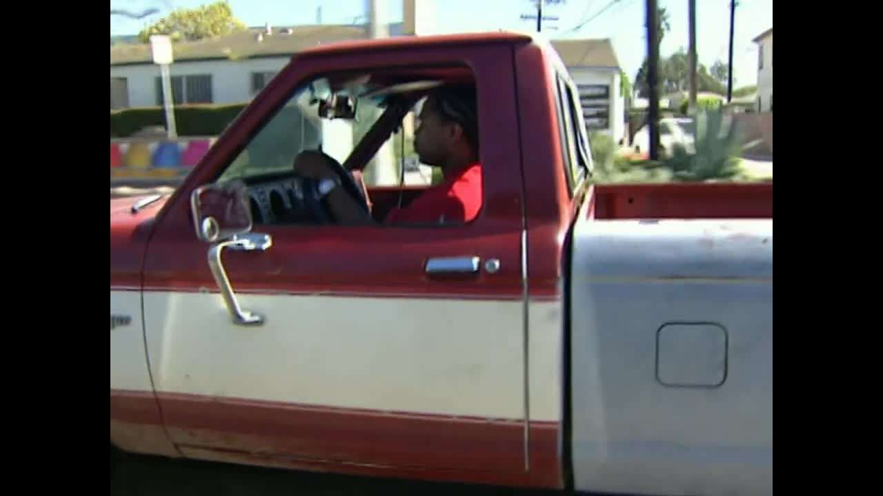 A red and white pickup truck rolls past, driven by a man in a red shirt. He's gripping the steering wheel, focused on the road ahead.
