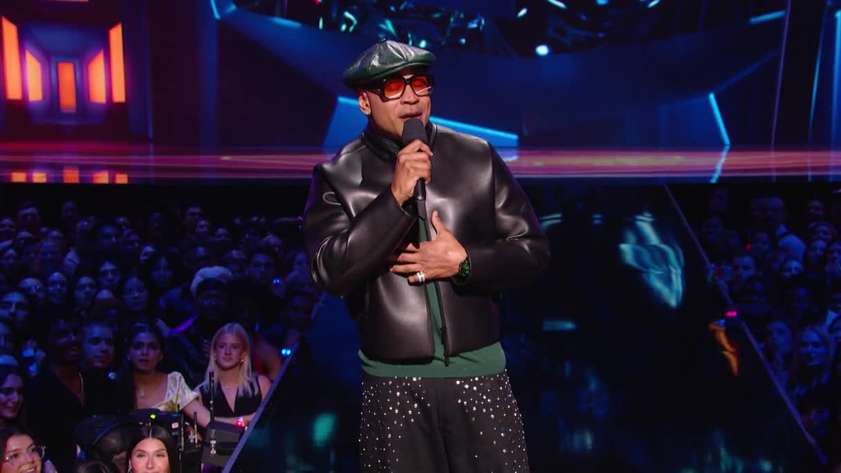 LL Cool J, in a leather jacket and cap, holds a microphone, addressing the crowd at an MTV MIAW awards show. The audience, packed close together, watches intently as he speaks.
