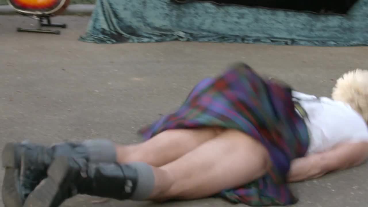 A person in a kilt and combat boots sprawls on the ground, their legs splayed. The kilt is lifted, revealing a glimpse of their skin.
