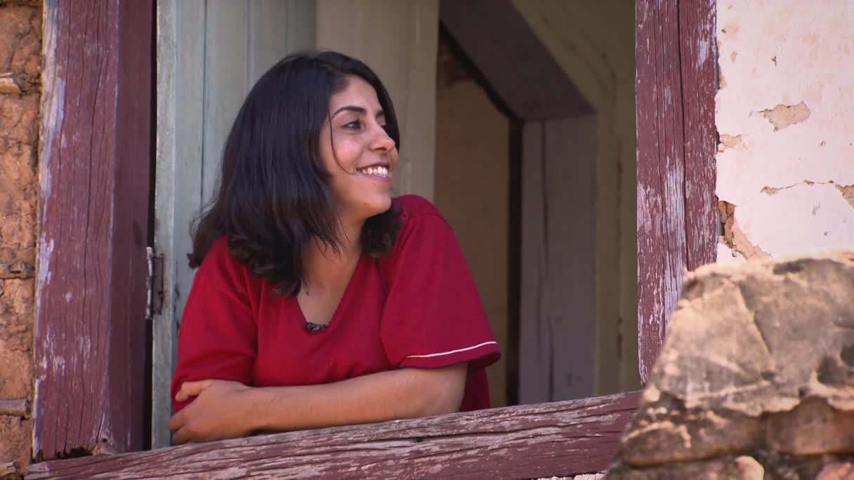 A woman in a red shirt leans on a weathered windowsill, a warm smile on her face. The scene feels like it's in Brazil, maybe for an MTV show, with the sunlight highlighting the peeling paint around the window frame.
