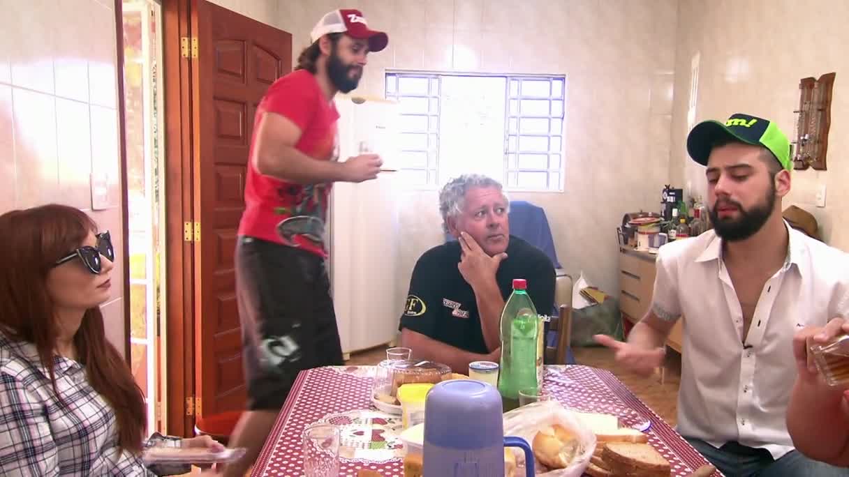 A man in a red shirt walks toward the table, holding a glass.  Two other men and a woman sit around a table covered in food, as if they're in a casual scene from a Brazilian MTV show.
