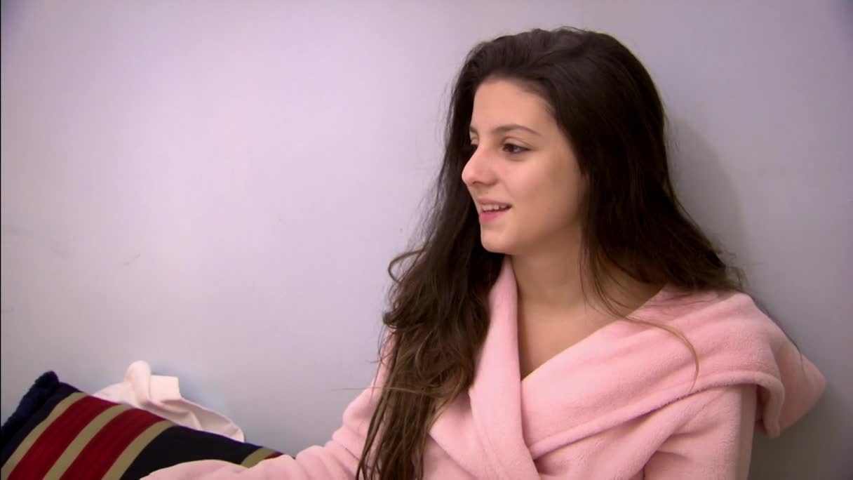 A young woman in a pink robe smiles, her dark hair cascading over her shoulders. She sits near a pillow with red and blue stripes, probably on a bed.
