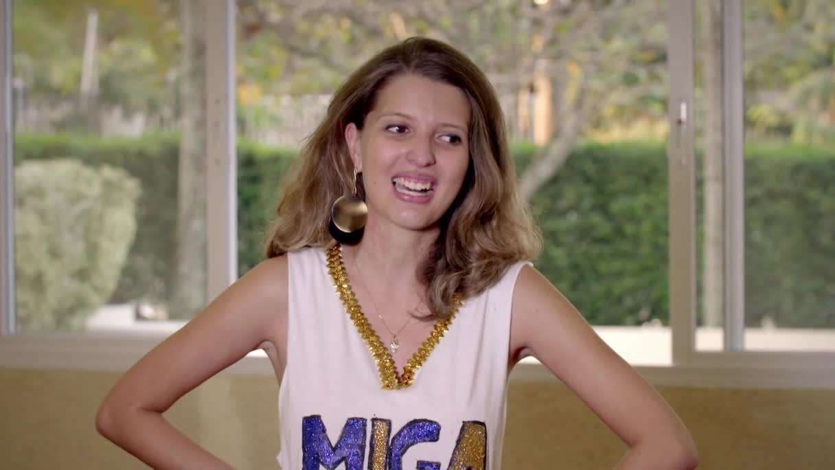 The woman, wearing a "MIGA" shirt, is smiling and gesturing with her hands. Sunlight streams through the window behind her as she speaks, probably on set for MTV Brazil.
