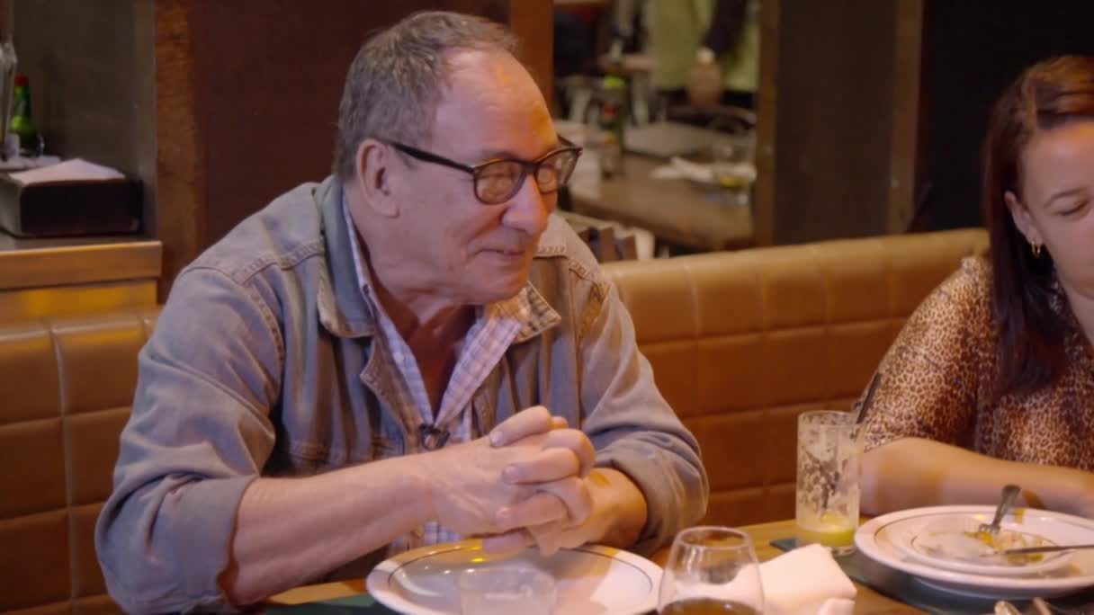 The man in glasses sits at a table, his hands clasped in front of him. He appears to be listening intently, a small smile playing on his lips.
