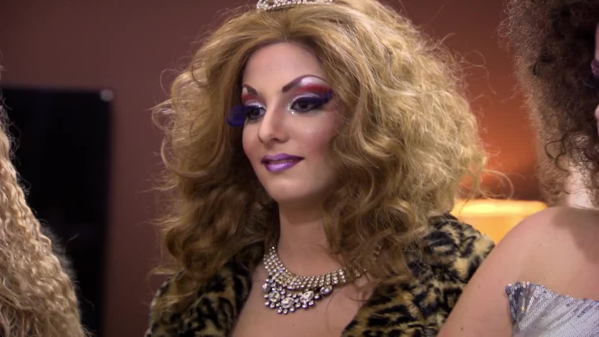 A contestant, decked out in full drag, is looking down, a tiara sparkling atop their voluminous, blonde wig. The camera seems to be focused on the intricate makeup and the shimmering necklace.
