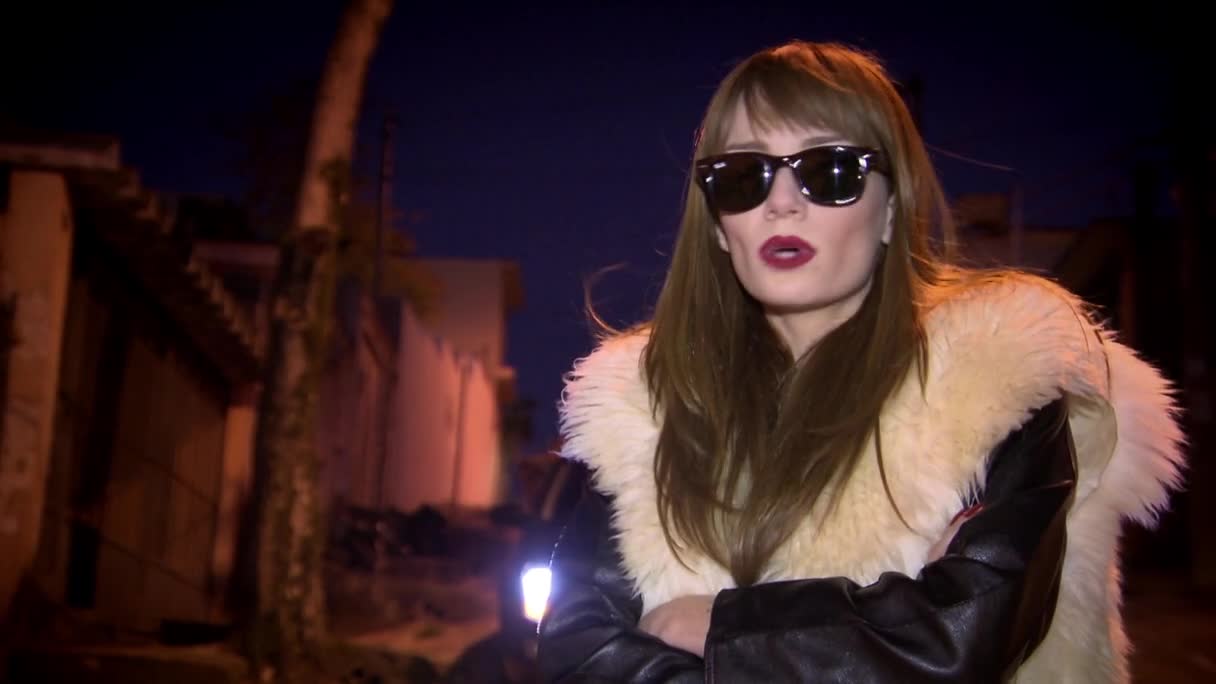 Under the night sky, a woman in sunglasses and a fur-trimmed jacket stands in front of a camera. The streetlights illuminate the scene as she speaks, her red lipstick a stark contrast to the dark backdrop.
