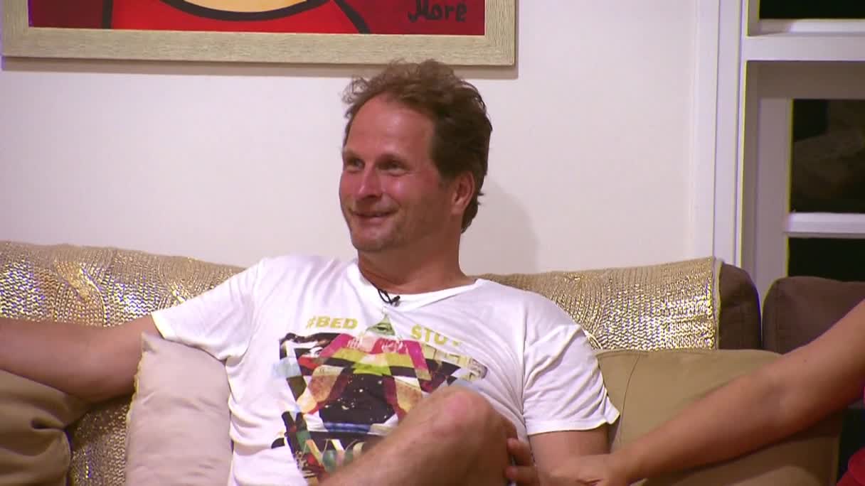 A man in a white t-shirt sits on a couch, smiling and looking to his right. The scene is lit by a warm, indoor light, and the couch has a decorative, gold-colored throw.
