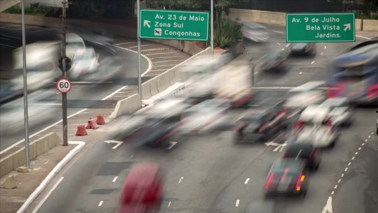 Cars streak along the highway, their shapes distorted by speed, a constant flow heading towards the Zona Sul. Above, the green street signs point towards different districts of São Paulo.
