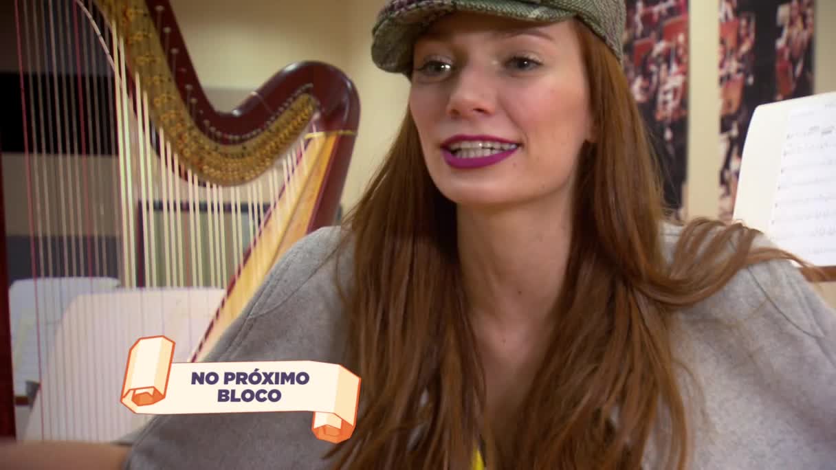 A woman with auburn hair and a green cap smiles, her lips painted a deep purple. Beside her, a harp's strings gleam, and a banner reads "NO PRÓXIMO BLOCO," indicating the next segment of the MTV Brazil broadcast.
