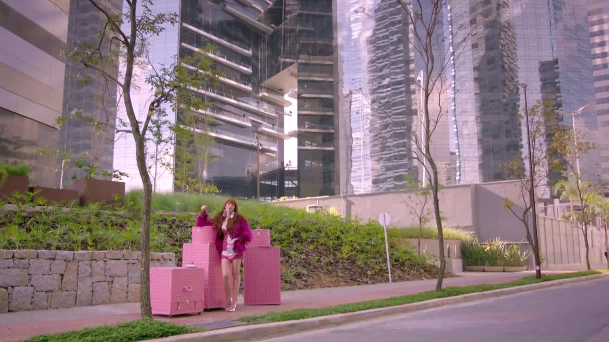 A woman in a bright pink furry jacket and matching shorts stands with a stack of pink luggage. She's on a sidewalk next to a street, with sleek, modern buildings reflecting the sky behind her.