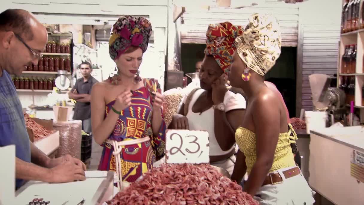 A vendor in Brazil hands a small item to a woman in a vibrant, patterned dress, her head wrapped in a colorful scarf. Behind them, two other women, also adorned with striking headwraps, engage in conversation at a market stall piled high with dried goods.