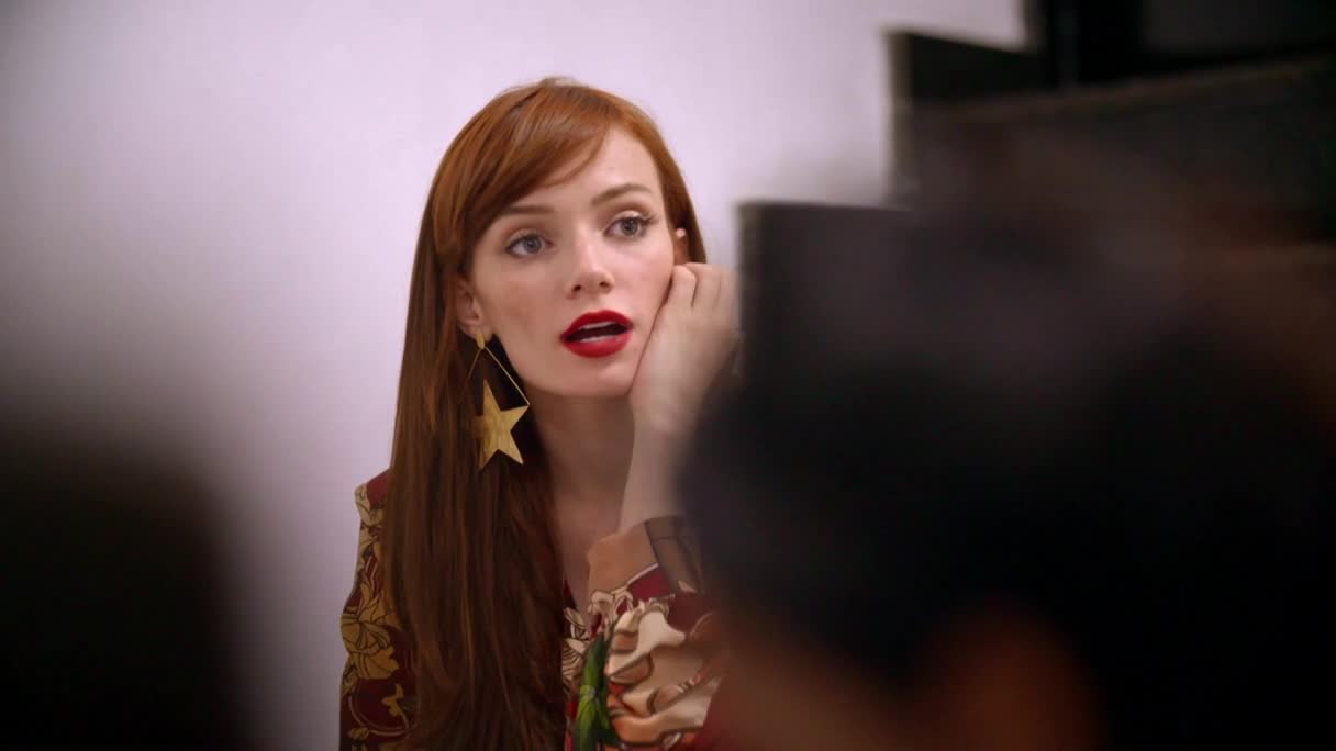 A woman with long auburn hair and bright red lipstick adjusts a large, golden star earring. She's wearing a patterned top, and her expression suggests she's mid-conversation.