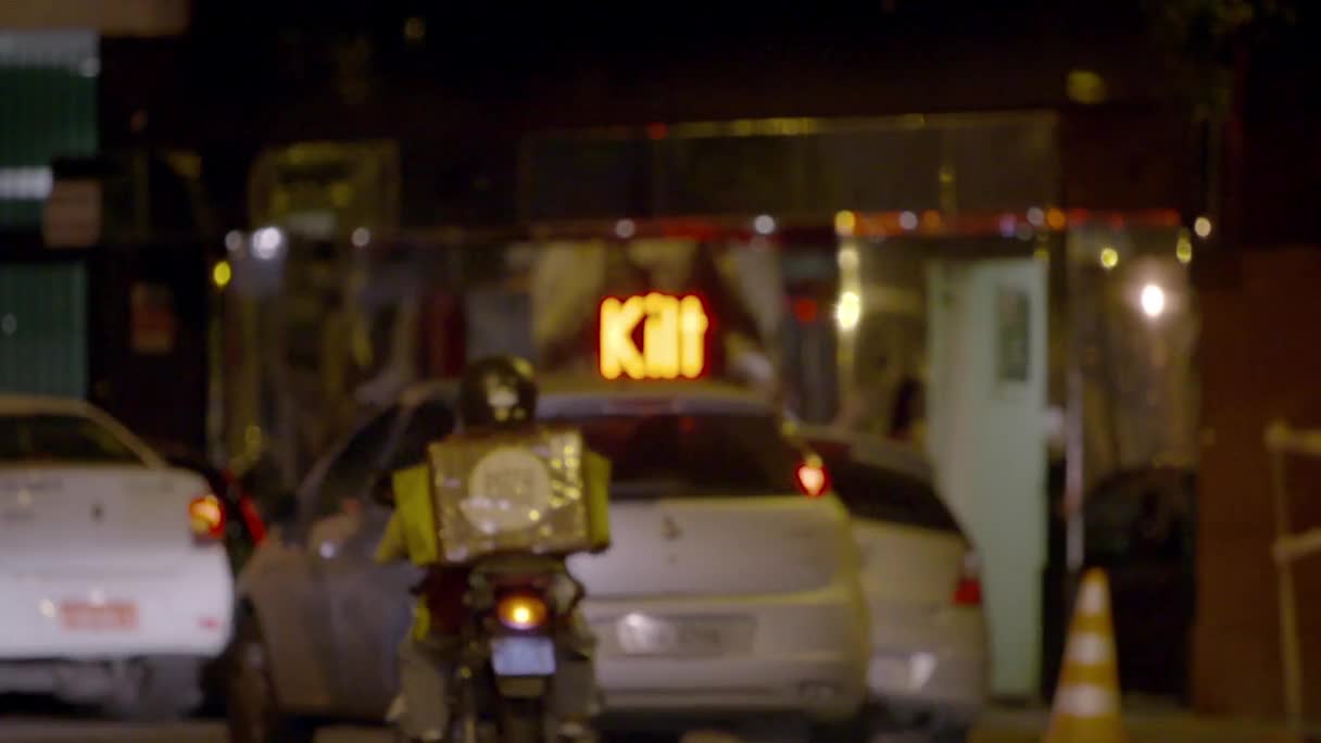 A motorcycle rider with a bright yellow delivery box navigates through traffic. Above them, a neon sign glows "Kitt" in orange letters.
