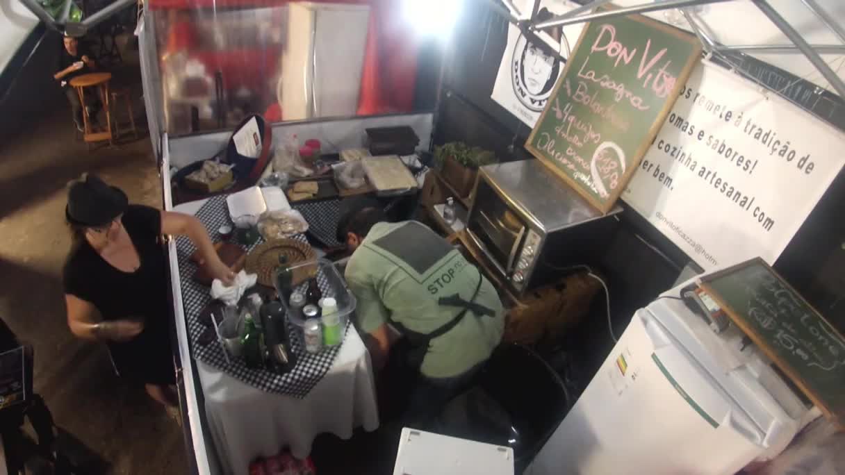 A woman in a black hat wipes down a checkered tablecloth at a food stall. Nearby, a person in a green apron bends over, working behind the counter.