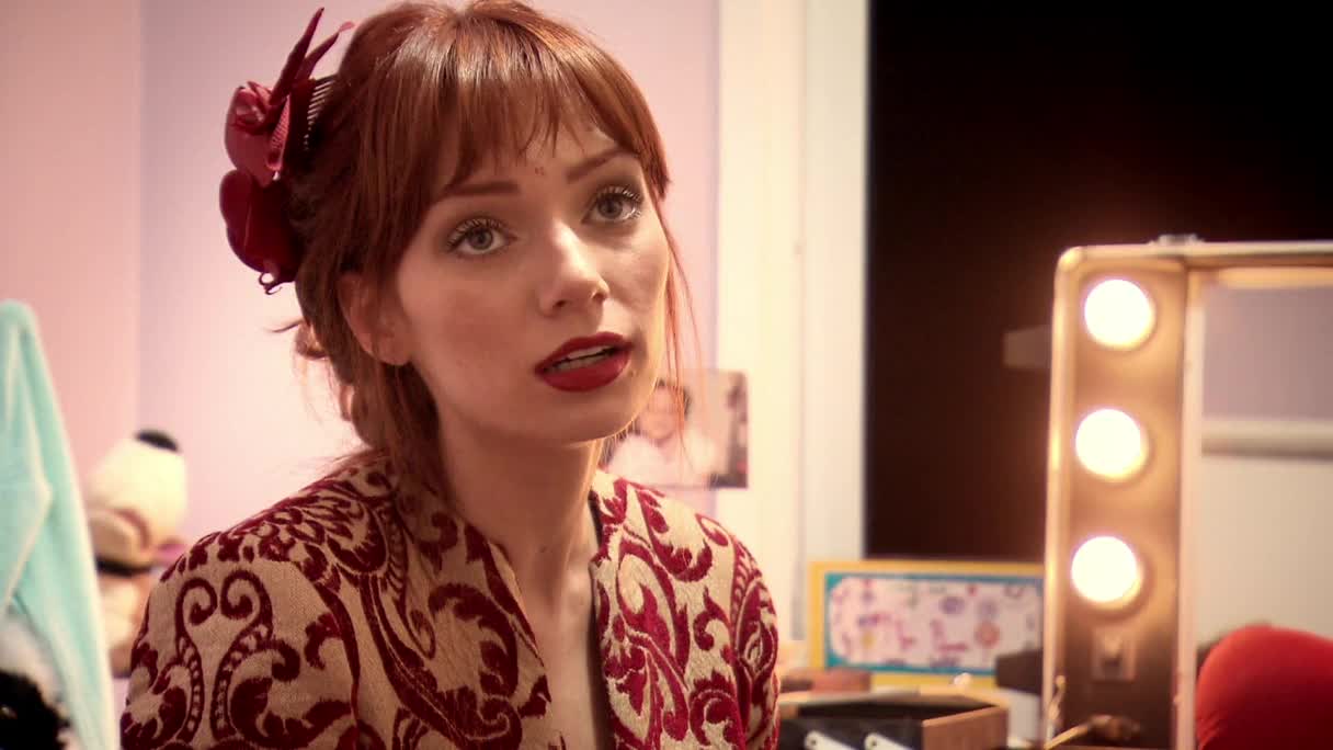 A woman with bright red lipstick and a large red flower in her auburn hair leans forward, her patterned jacket catching the light. A makeup mirror with illuminated bulbs sits to her right, reflecting a dimly lit room.
