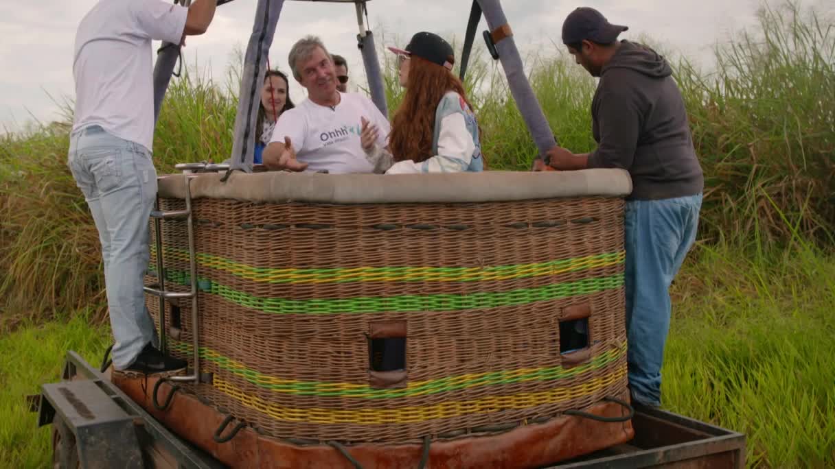 The hot air balloon basket is ready for launch, with people inside and a crew member standing on the edge. This looks like part of a segment for MTV Brazil, perhaps a travel show. The hot air balloon basket is ready for launch, with people inside and a crew member standing on the edge. This looks like part of a segment for MTV Brazil, perhaps a travel show.