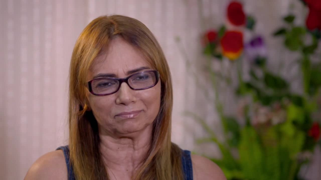 A woman with auburn hair and glasses looks directly ahead, a faint smile on her lips. Behind her, a blurred arrangement of red and yellow flowers adds a touch of color to the scene. A woman with auburn hair and glasses looks directly ahead, a faint smile on her lips. Behind her, a blurred arrangement of red and yellow flowers adds a touch of color to the scene.