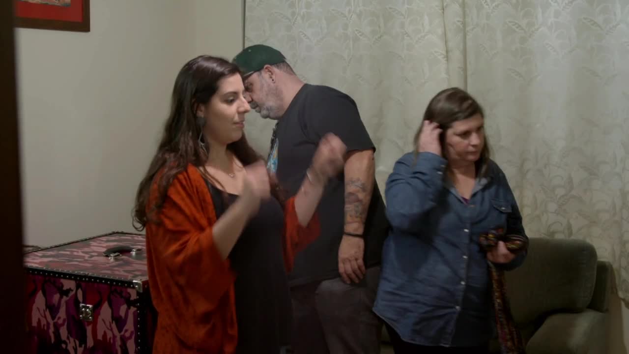 A young woman in a bright orange shawl is talking with a man wearing a green cap. To their right, another woman adjusts her hair, clutching a patterned scarf. A young woman in a bright orange shawl is talking with a man wearing a green cap. To their right, another woman adjusts her hair, clutching a patterned scarf.