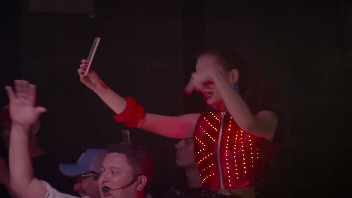 A performer in a red, light-up vest holds up a phone, capturing the energy of the crowd in Brazil. People around her wave and cheer, caught up in the excitement of the MTV event. A performer in a red, light-up vest holds up a phone, capturing the energy of the crowd in Brazil. People around her wave and cheer, caught up in the excitement of the MTV event.