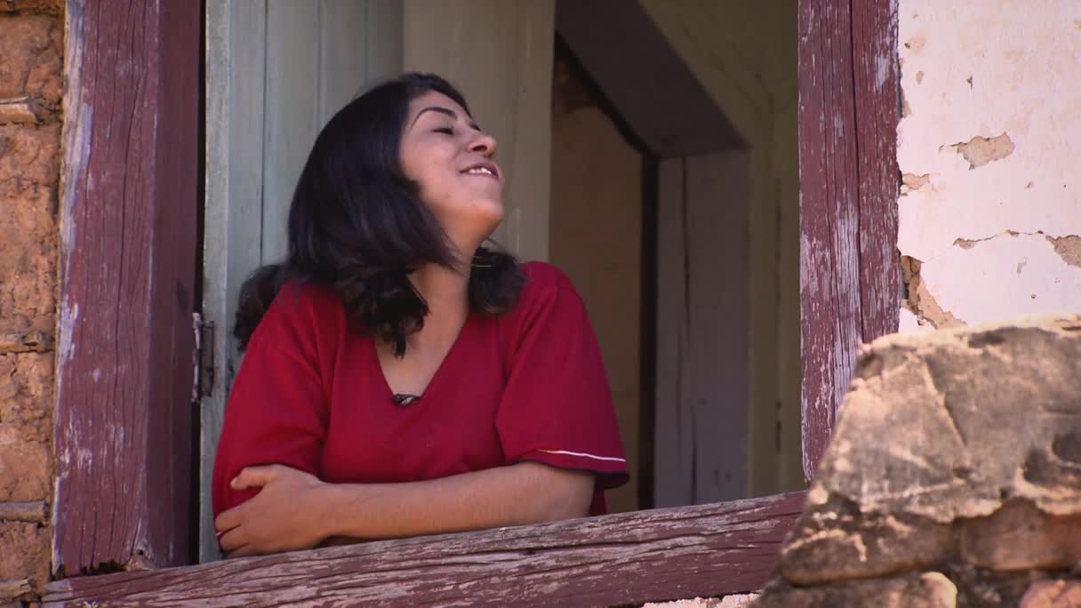 A young woman in a red shirt leans out of a weathered window frame, her face tilted up towards the sun. The rough, sun-baked walls of her home in Brazil are visible behind her.