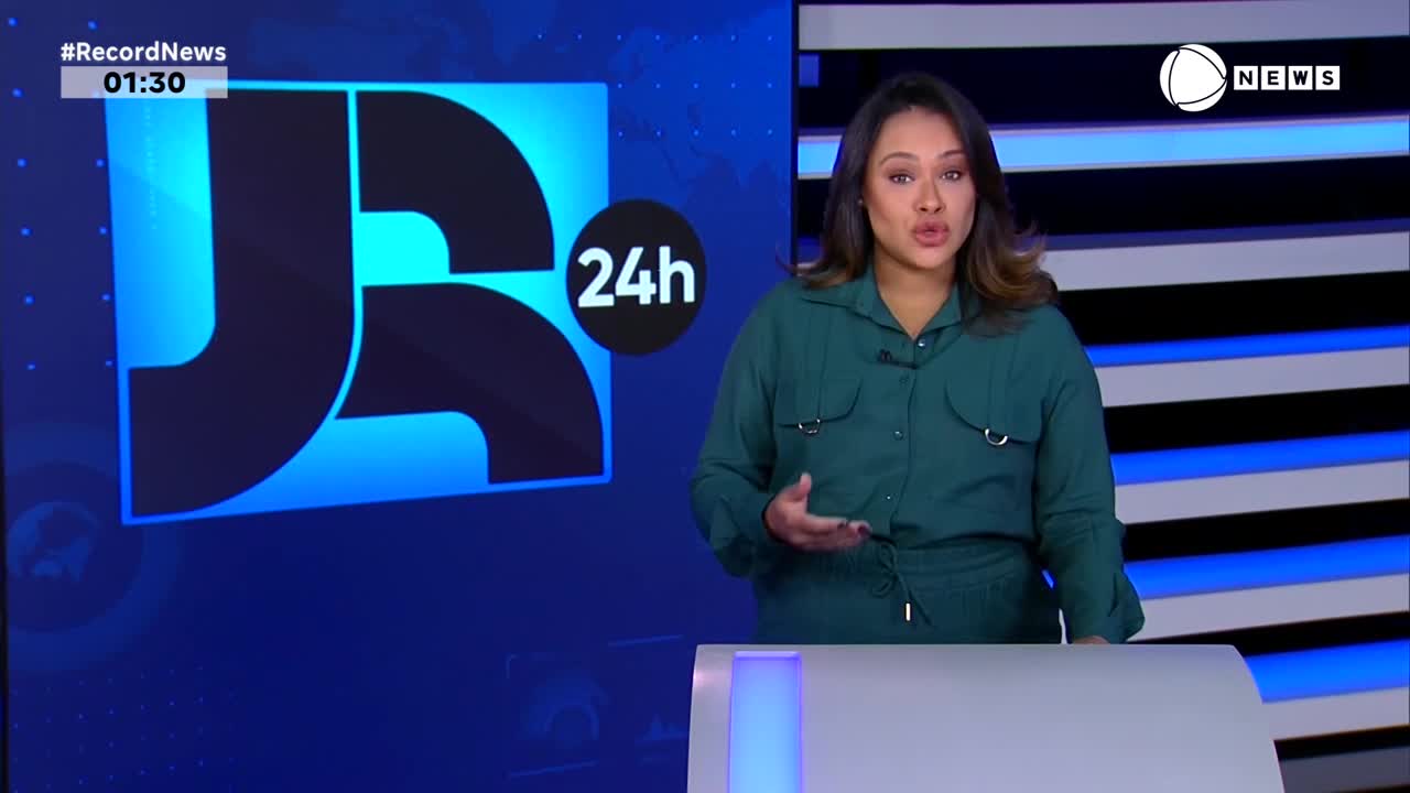 The Record News anchor, in a green top, gestures with her hands as she speaks. Behind her, the JR logo and a "24h" graphic are displayed on a blue backdrop.
