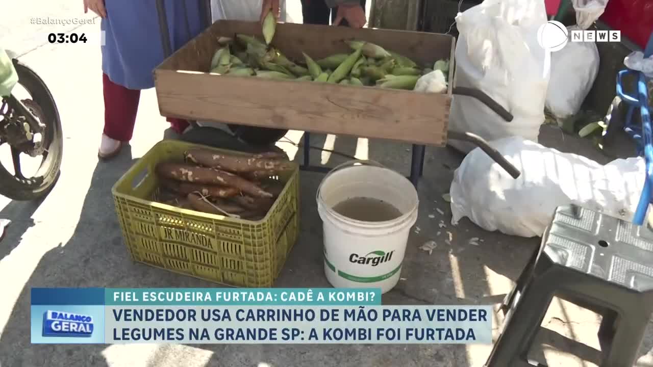 A vendor in Brazil, as reported by Record News, is selling vegetables from a cart. Corn and cassava are displayed in wooden and plastic crates, with a bucket of water nearby.
