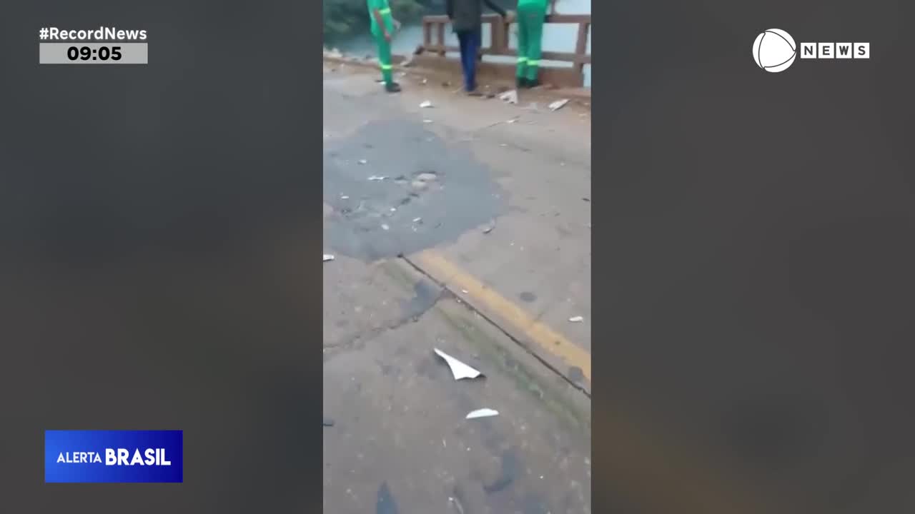 On this bridge, workers in green coveralls stand near a railing.  Paper scraps litter the wet pavement as the Record News broadcast continues.
