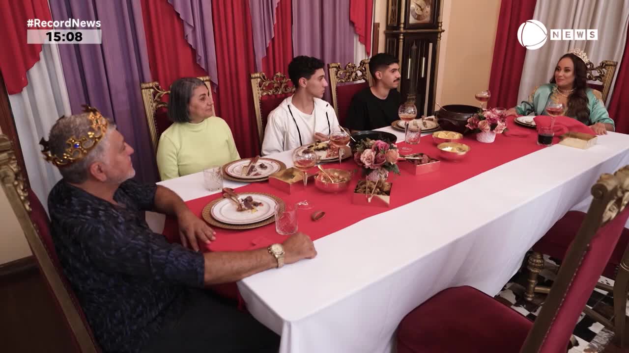 A family, seated around a long table, is gathered for a meal in a room decorated with red and purple drapes. The man at the head of the table wears a crown, and they're likely being filmed for a segment on Record News in Brazil.
