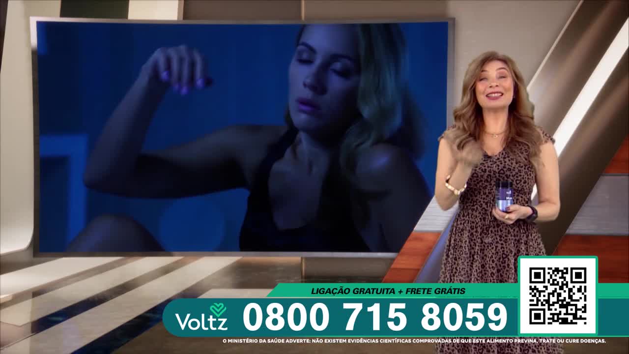On a Brazil Record News set, a woman in a leopard-print dress holds a small bottle while smiling at the camera. Behind her, a screen shows a woman in a dark room, her arm raised.

