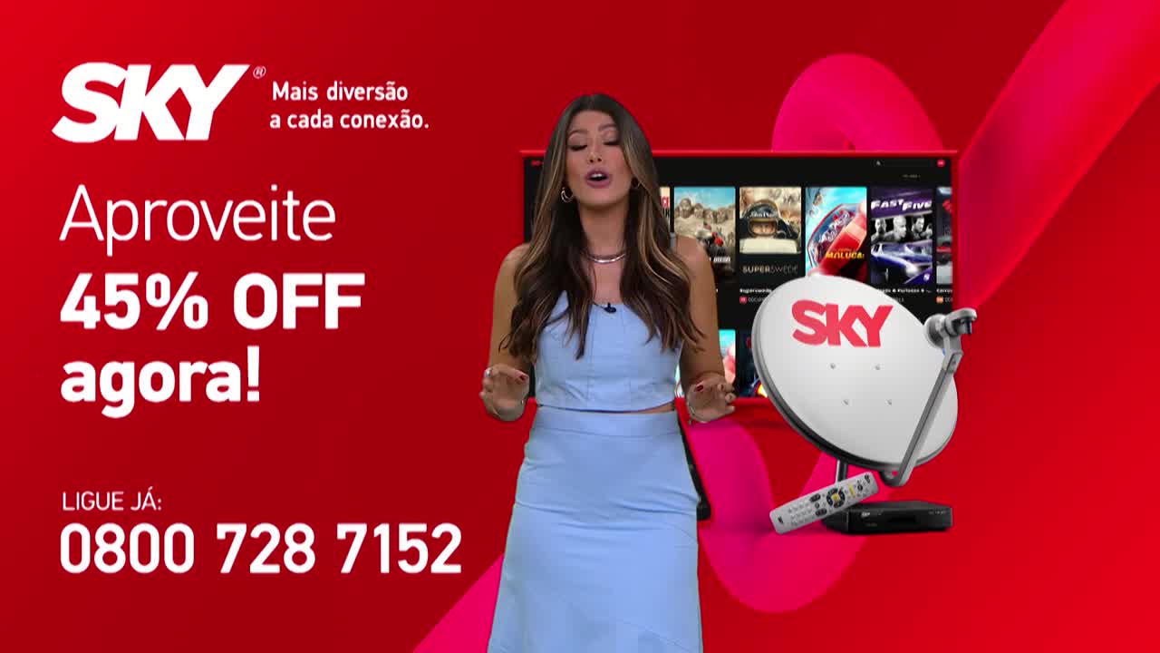 A woman in a light blue outfit stands in front of a red backdrop, gesturing with her hands.  Behind her, a TV screen and a satellite dish with the SKY logo are prominently displayed.
