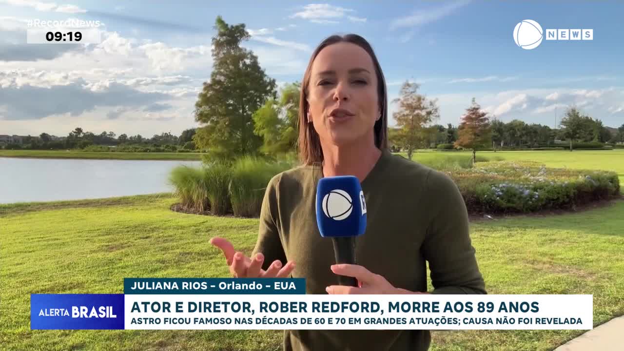 A woman in a green shirt speaks into a microphone, gesturing with her hands in front of a lake and green space. The Record News logo is visible on the microphone, and the bottom of the screen displays a headline about Robert Redford's death.
