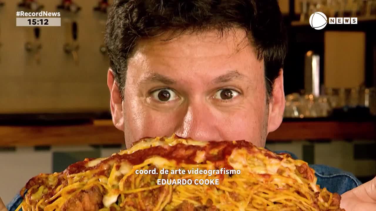 A man's wide-eyed expression peers over a massive plate of spaghetti, the food obscuring most of his face. The Record News logo and time stamp are visible in the corner.
