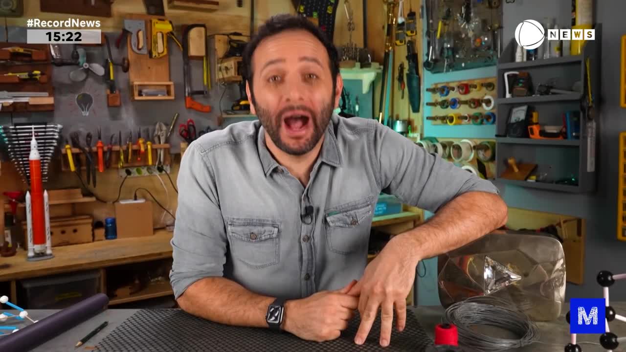 On the set of Record News in Brazil, a man in a denim shirt gestures excitedly with his hands. He's sitting at a workbench, with a silver award and various crafting materials in front of him.
