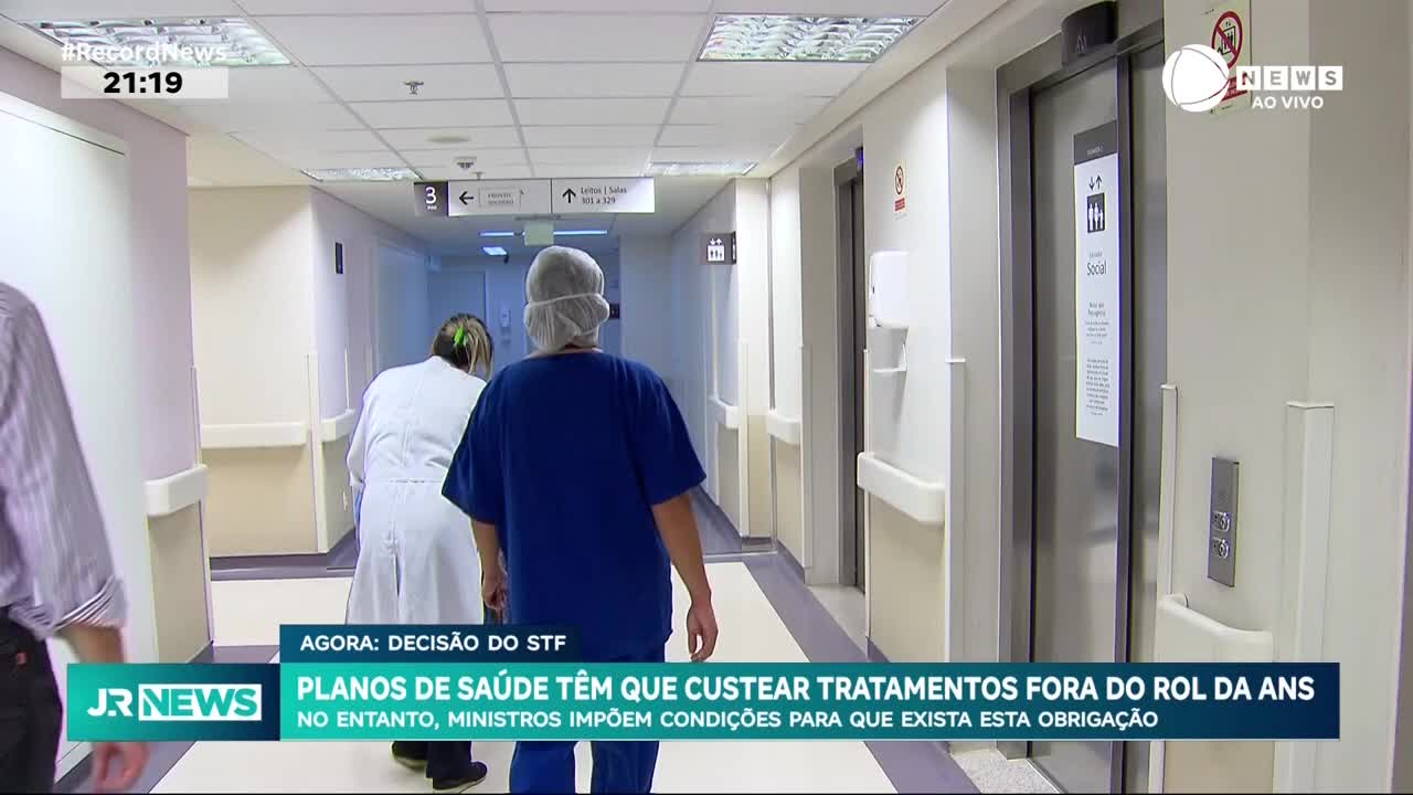 Two medical professionals in scrubs and a lab coat walk down a hospital corridor. A broadcast from Brazil's Record News is reporting on a Supreme Court decision regarding health insurance coverage.
