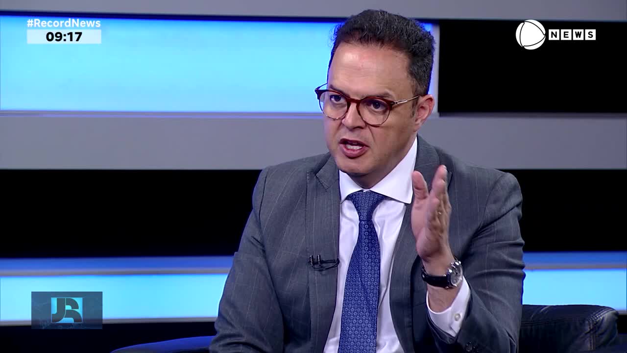 The man in the suit, wearing glasses, gestures with his right hand as he speaks on Record News. The time, 09:17, is displayed in the upper left corner.
