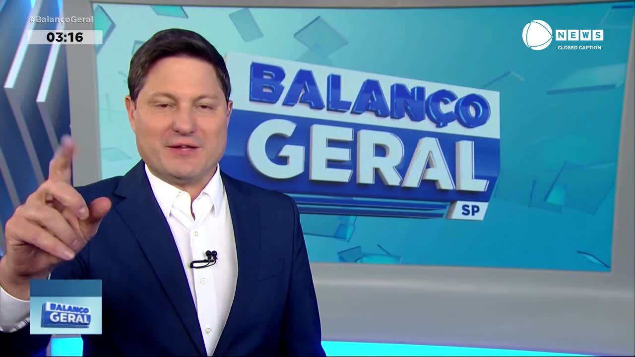 The man in the blue suit is gesturing with his right index finger as he speaks. Behind him, the "Balanço Geral" logo and the Record News logo are clearly visible on the screen.
