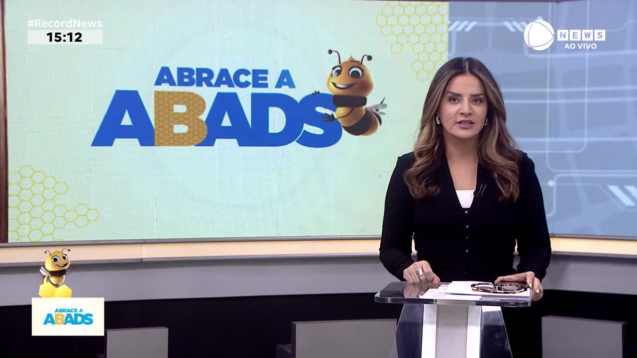 The Record News presenter stands behind a clear desk, speaking directly to the camera. Behind her, a graphic with the words "ABRACE A ABADS" and a cartoon bee fills the screen.
