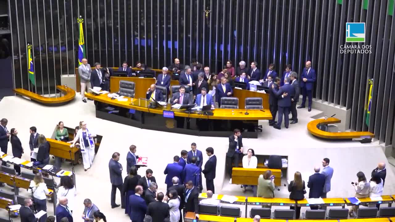 In the Brazilian Chamber of Deputies, people in suits mill about, some in small groups, others standing alone. A few men in the center are embracing, while others seem to be talking animatedly.
