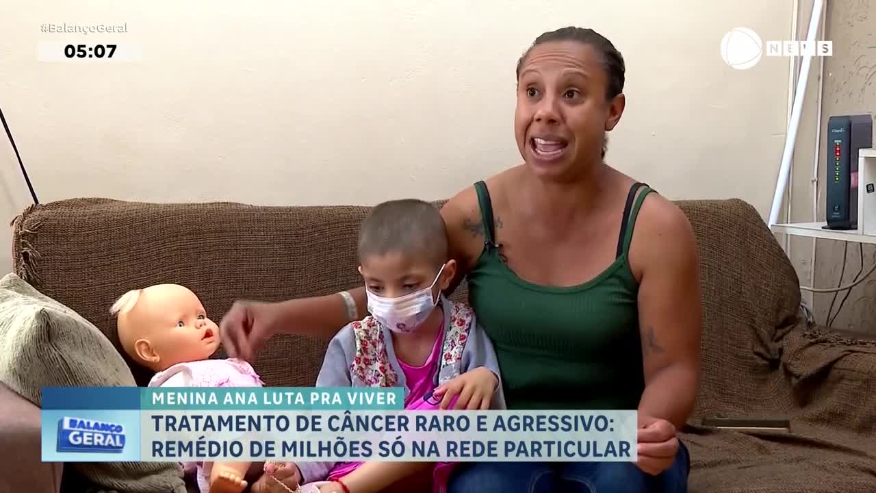 A woman speaks animatedly while sitting next to a young girl wearing a mask and a doll. The text overlay from Record News discusses a girl named Ana fighting for her life against a rare cancer.