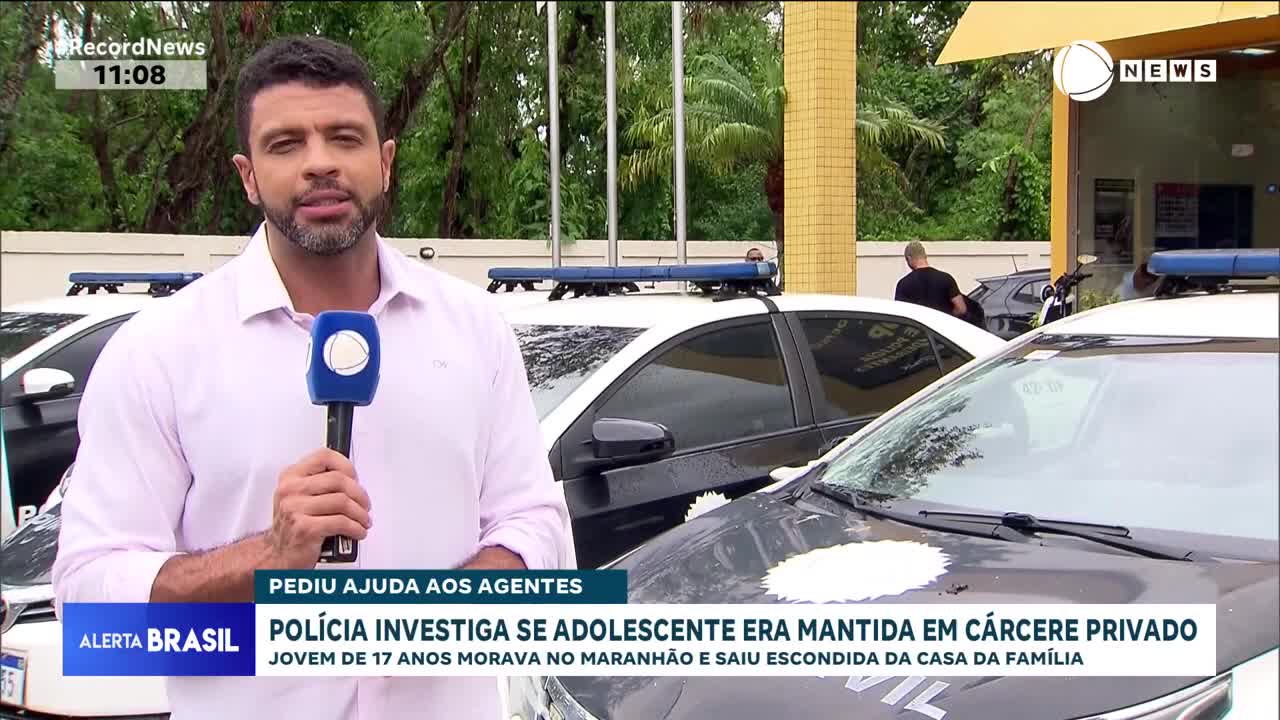 A reporter stands in front of several police cars, holding a microphone. In the background, a man walks away from the building.