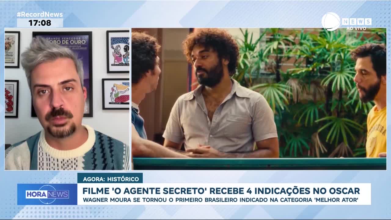A man with a beard and curly hair, wearing a light brown shirt, looks intently at another man. Behind them, lush green foliage frames the scene.