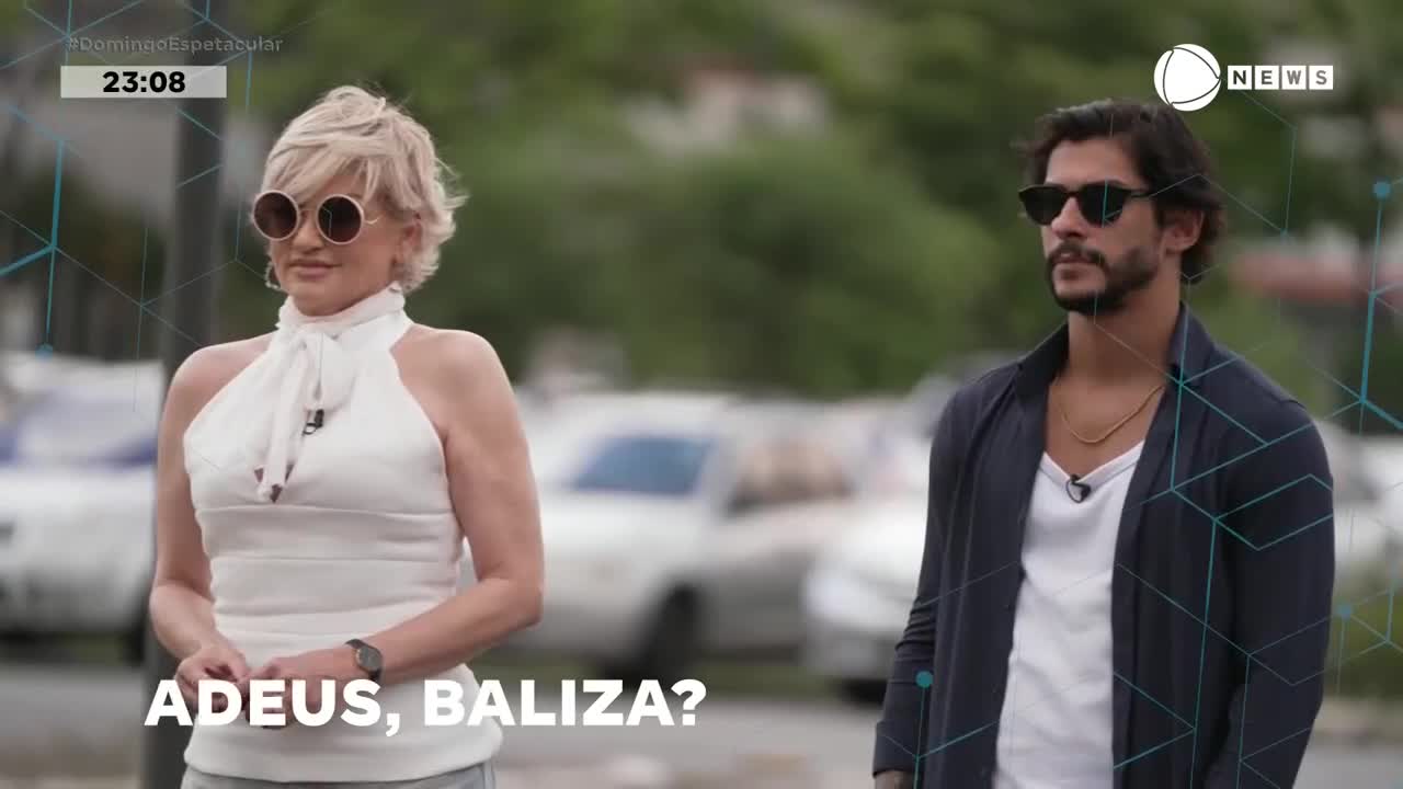 A woman with blonde hair and large sunglasses stands still, her white halter top catching the light. Beside her, a man with dark hair and sunglasses walks away, his open navy cardigan revealing a white t-shirt.