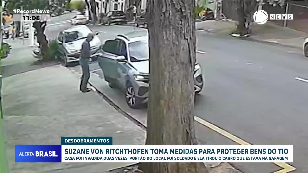 A man in a dark jacket and jeans stands by the open door of a silver SUV. He appears to be looking at something on the sidewalk. The car is parked on a street in Brazil, with a large tree trunk obscuring part of the view.